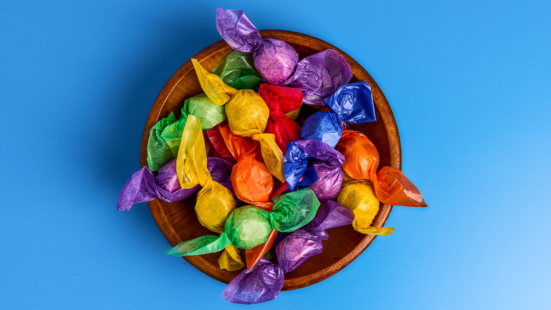 Wooden bowl filled with colorful candy wraps on a blue background